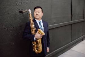 An image of Dr. Jordan VanHemert holding a saxphone and leaning against a wall